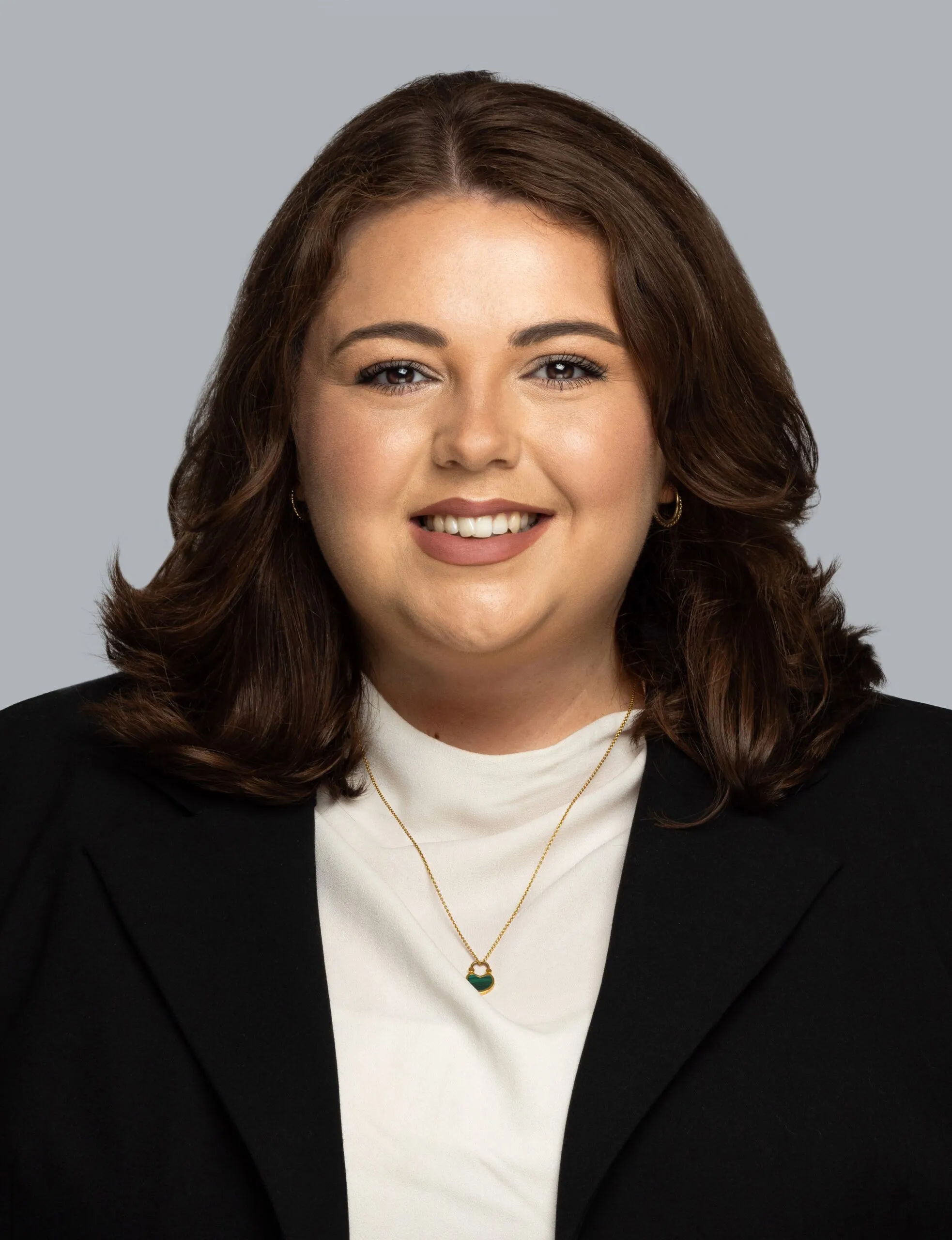 Abigail Clements-Bewley wears a black jacket white top and gold chain necklave. Her dark brown hair is shoulder-length and she is smiling at the camera.