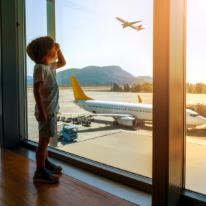 Boy about age 5 in tshirts, shorts and trainers standing at airport window looking at aeroplanes. There's an airplane in the background taking off and another parked on runway.