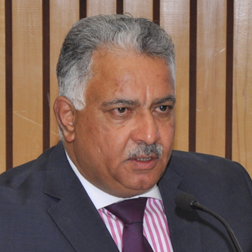 Anil Malhotra is wearing a dark grey suit jacket, pink and white striped collared shirt, and a burgundy tie. His hair is short and he has a moustache. He's speaking into a podium mic.