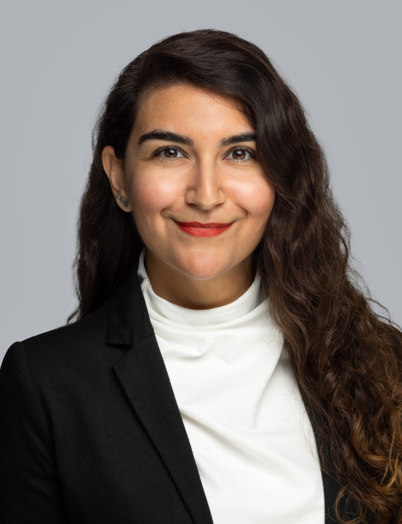 Sherry Georgaki has long wavy hair, and is smiling at the camera. She wears a black blazer and white top.