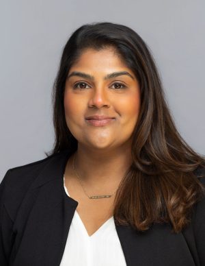 Geeta's long, dark wavy hair is swept over her left shoulder. She's smiling at the camera, and wears a black blazer, white top, and a gold necklace.