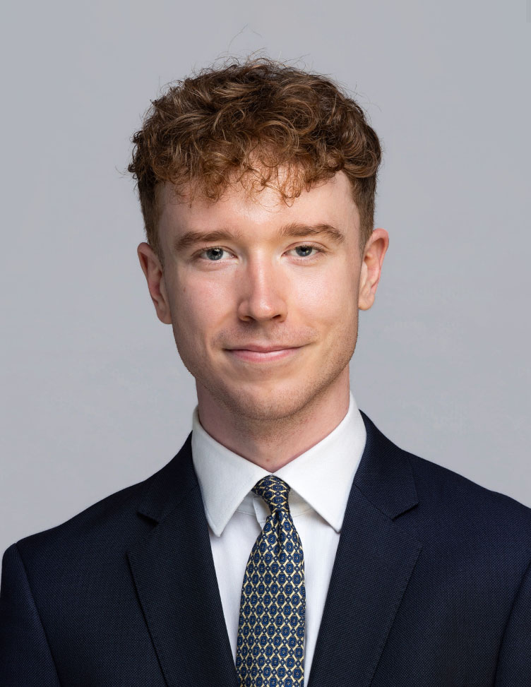 Owen wears a dark suit, white shirt and patterned grey tie. He's gently smiling at the camera.